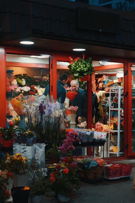 The image depicts the exterior of a flower shop at dusk with a vibrant display of fresh flowers arranged outside, including colorful bouquets and potted plants. Through the large glass window, a varie