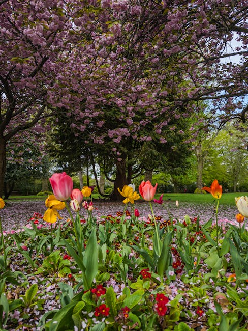 A vibrant garden scene featuring a variety of freshly arranged flowers in the foreground, including pink tulips, yellow pansies, and red primroses, all with lush green stems and leaves. These flowers 
