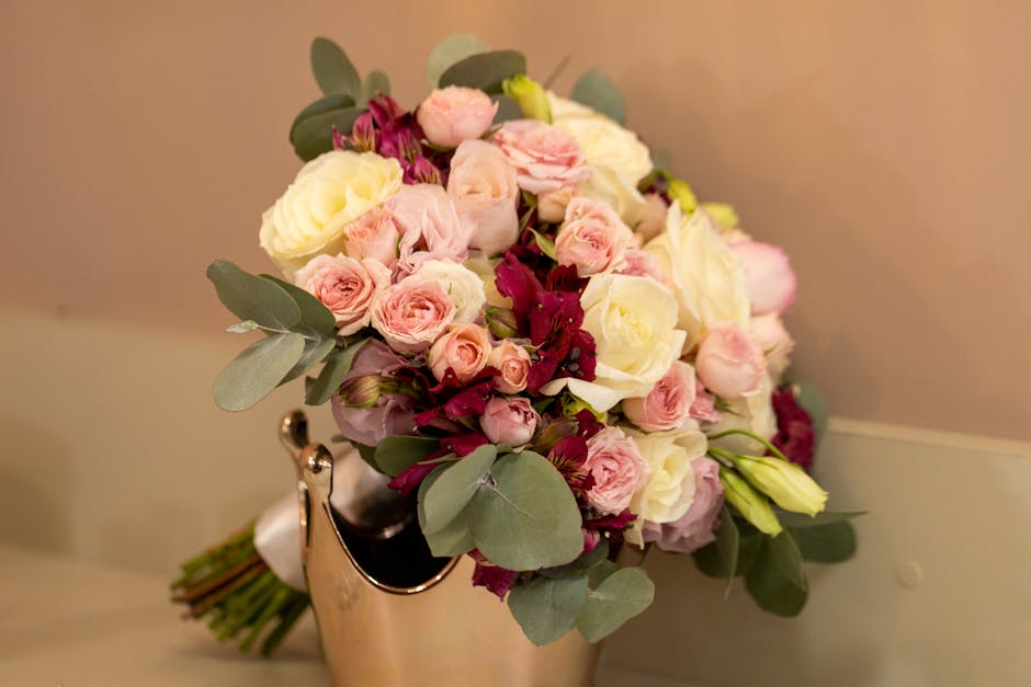 A floral bouquet featuring a combination of soft pink and creamy white roses, complemented by deep burgundy flowers and lush eucalyptus leaves. The arrangement is placed in a silver vase on a neutral-