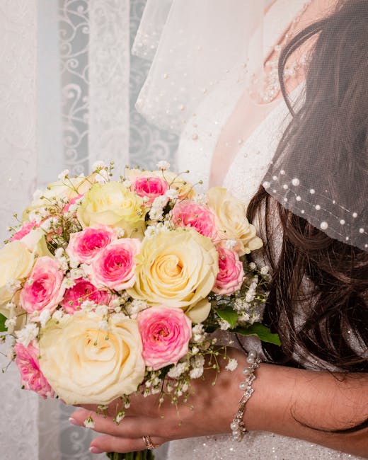 A close-up of a bride holding a floral bouquet with a mix of large, creamy white roses and smaller pink and white roses, complemented by sprigs of baby's breath. The bouquet is arranged in a rounded, 