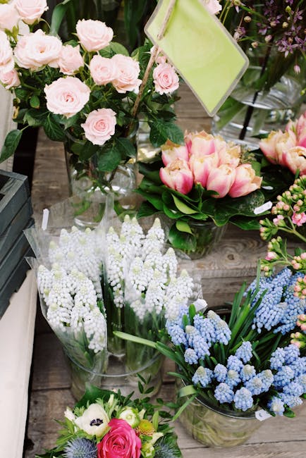 An assortment of fresh floral arrangements displayed on a rustic wooden surface, including a bouquet of pale pink roses with lush green foliage arranged in a clear glass vase, and a separate arrangeme